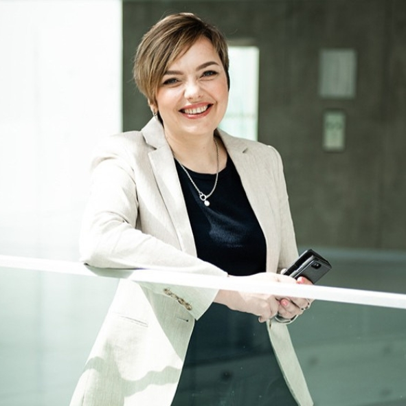 Laiminga moteris trumpais šviesiai rudais plaukais, vilkinti pilkšvos spalvos švarką ant juodos palaidinės, laikanti išmanųjį telefoną ir besiremianti į stiklinį turėklą šviesiame moderniame interjere.